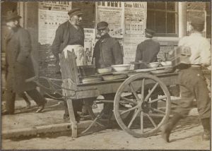 A pickle seller on an Amsterdam street in 1899. Behind him, a wall of layered bills, one of them announcing Circus Arena.