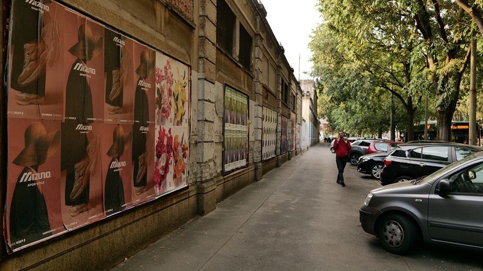 A long municipal wall in Milan pasted with a repeating Mizuno campaign, with framed notice panels further along the same wall and parked cars on the street.