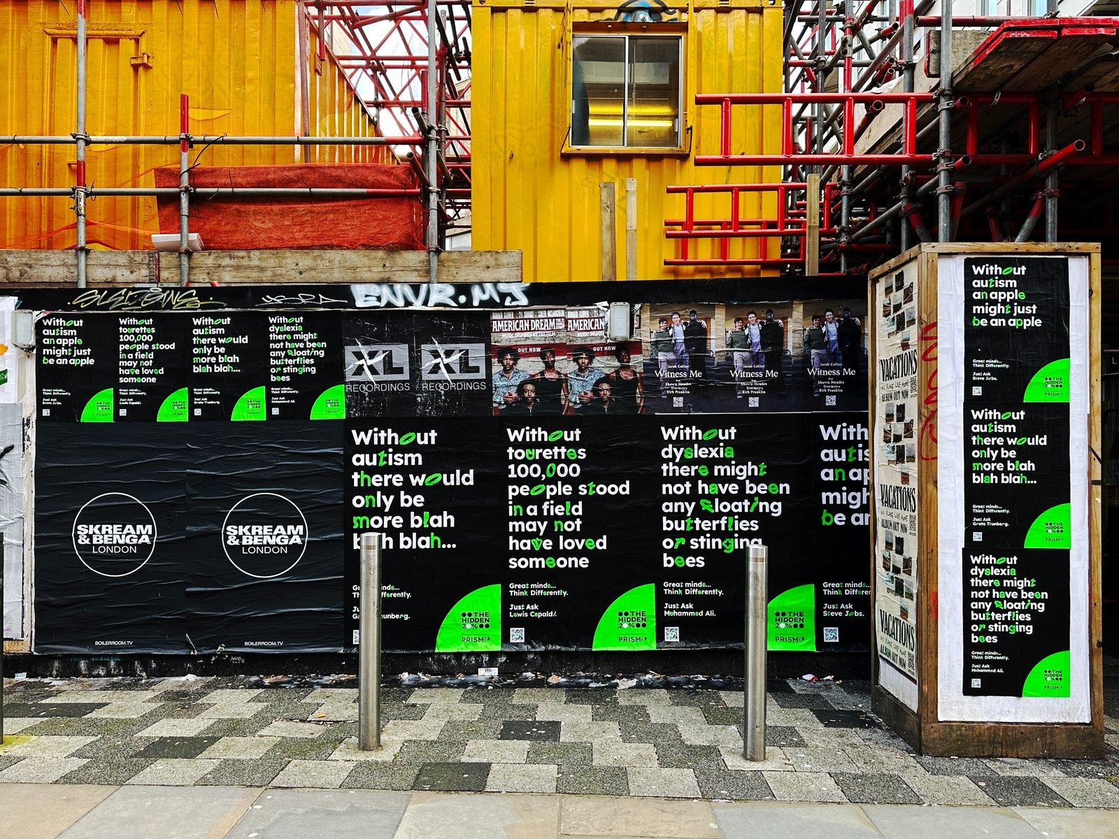 A construction hoarding in London pasted with overlapping campaigns from unconnected advertisers, several printed over each other, with graffiti tags across the top and scaffolding above.