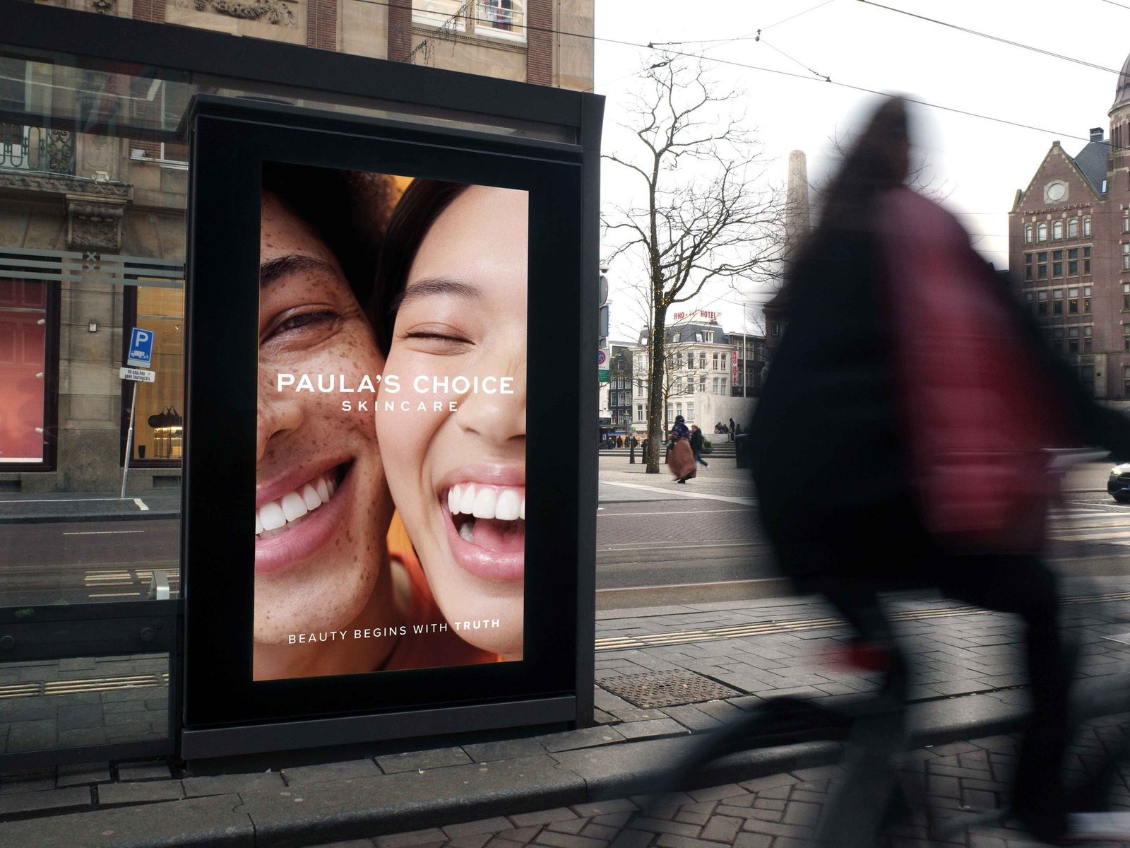 A backlit digital advertising panel built into a tram shelter in central Amsterdam, carrying a skincare campaign, with a cyclist passing in front of it.
