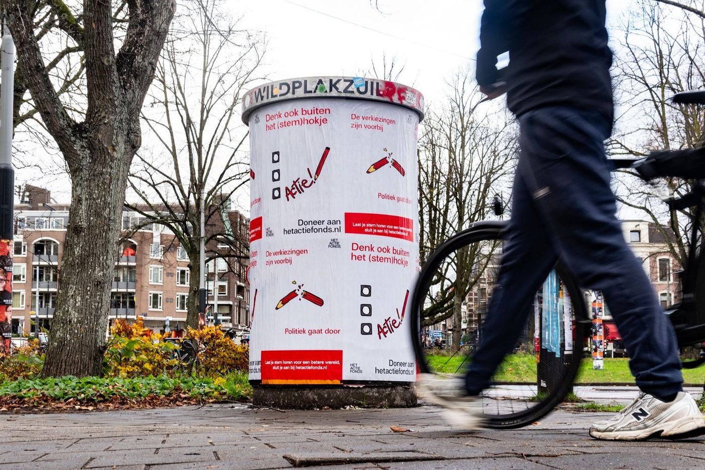 A cylindrical free posting column at the edge of a park in winter, pasted with a red and white campaign by a political fundraising foundation, a cyclist passing in the foreground.
