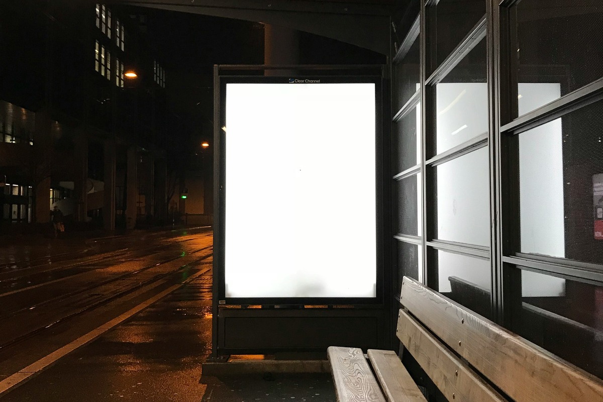 A tram shelter in Zurich at night. The backlit advertising panel is lit but completely empty, with the operator's mark across its head. Wet tram tracks run away to the left and a wooden bench to the right.